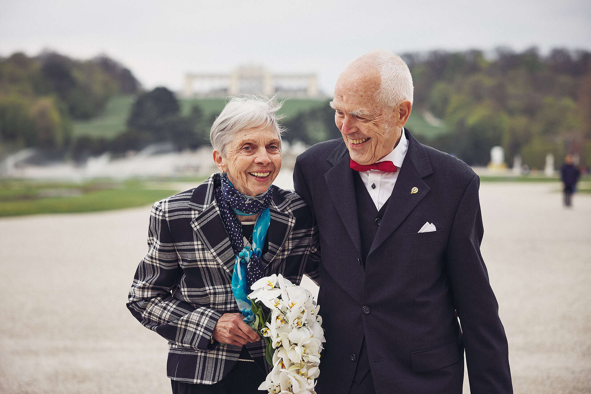 Älteres Brautpaar steht im Schlosspark, hinter ihnen sieht man die Gloriette. Die beiden lächeln und die Braut hält einen weißen Blumenstrauß in der Hand.